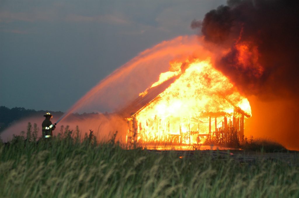 Ein Haus in Vollbrand, davor ein Feuerwehrmann, löschend
